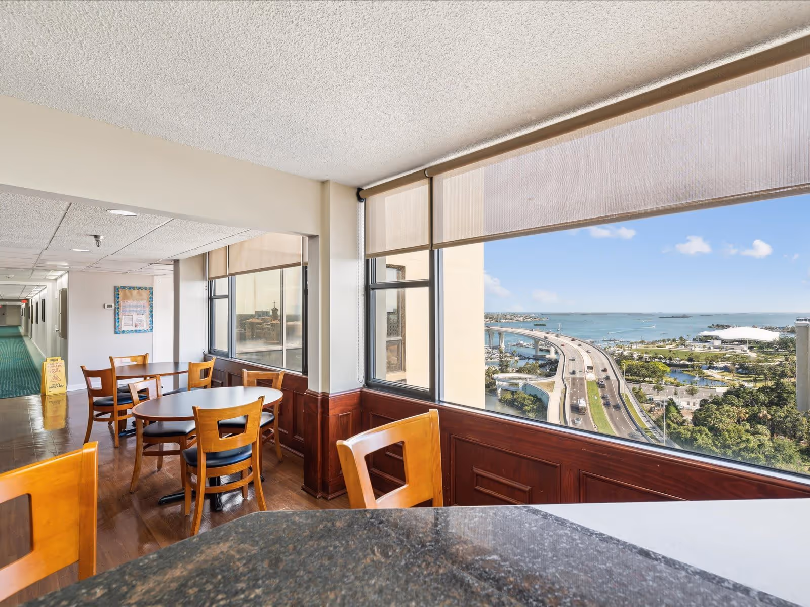 Interior view of a senior living facility dining area with wooden chairs and tables near large windows overlooking a highway, green trees, and a body of water under a blue sky with some clouds.