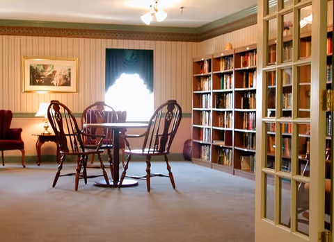 Cozy common room with a round table and wooden chairs, bookshelves, and armchairs by a curtained window.