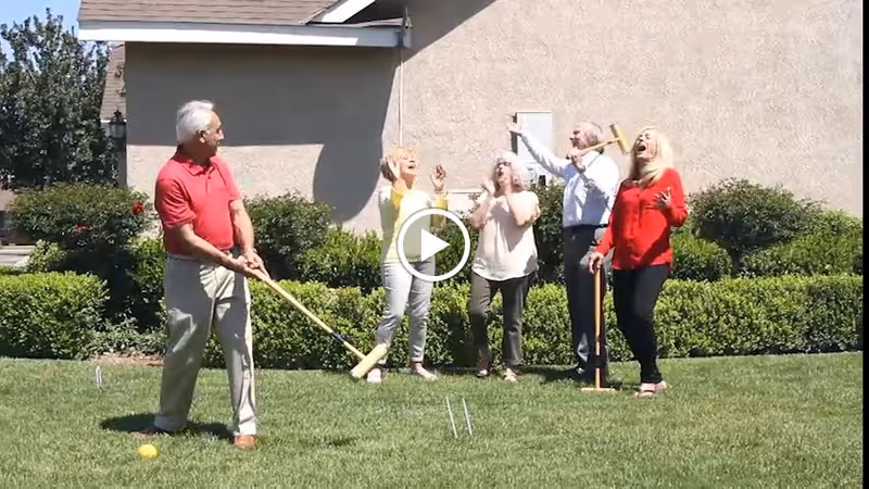A group of seniors playing croquet on a lawn in front of a building, with one man swinging a mallet while others cheer.