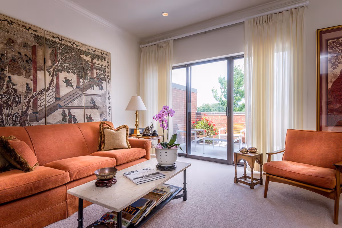 A cozy living room with an orange sofa and matching armchair, a marble coffee table with magazines and a potted orchid, a side table with a lamp and decorative items, and a large sliding glass door leading to a small outdoor patio with chairs and plants. The room features light-colored walls and carpet, with a large decorative wall hanging behind the sofa.