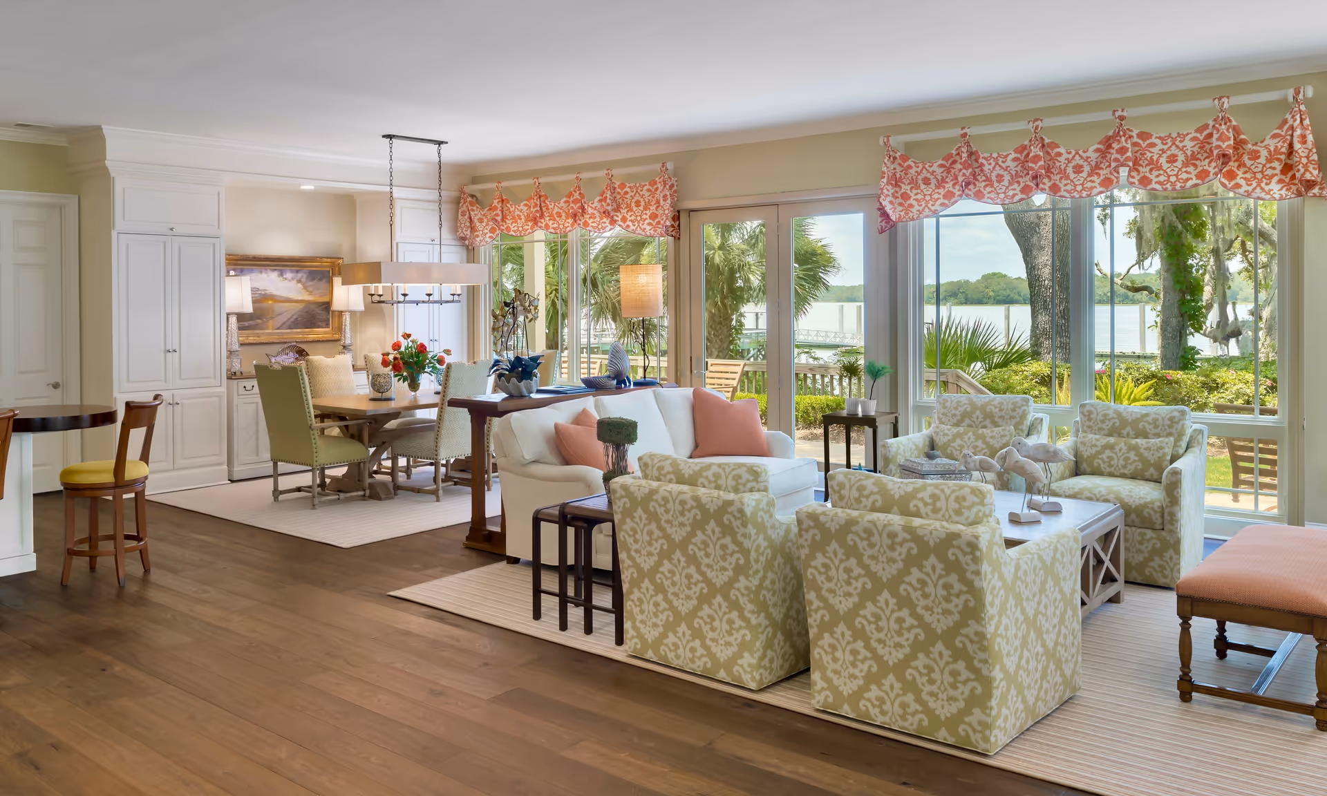 Open living and dining room with patterned armchairs, a sofa, dining table, and large windows overlooking a waterfront view.