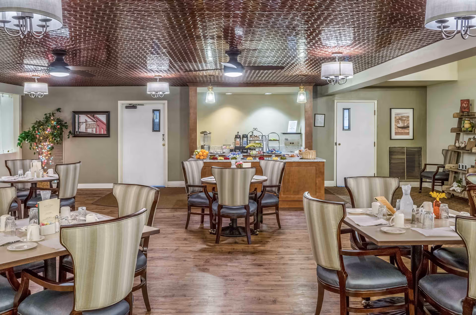 A dining room in a senior living facility with several tables and chairs arranged neatly. The tables are set with plates, cups, and utensils. In the background, there is a buffet area with fruit, beverages, and other food items. The room has a decorative ceiling with multiple light fixtures and ceiling fans. There are framed pictures on the walls and a small plant with string lights in the corner.
