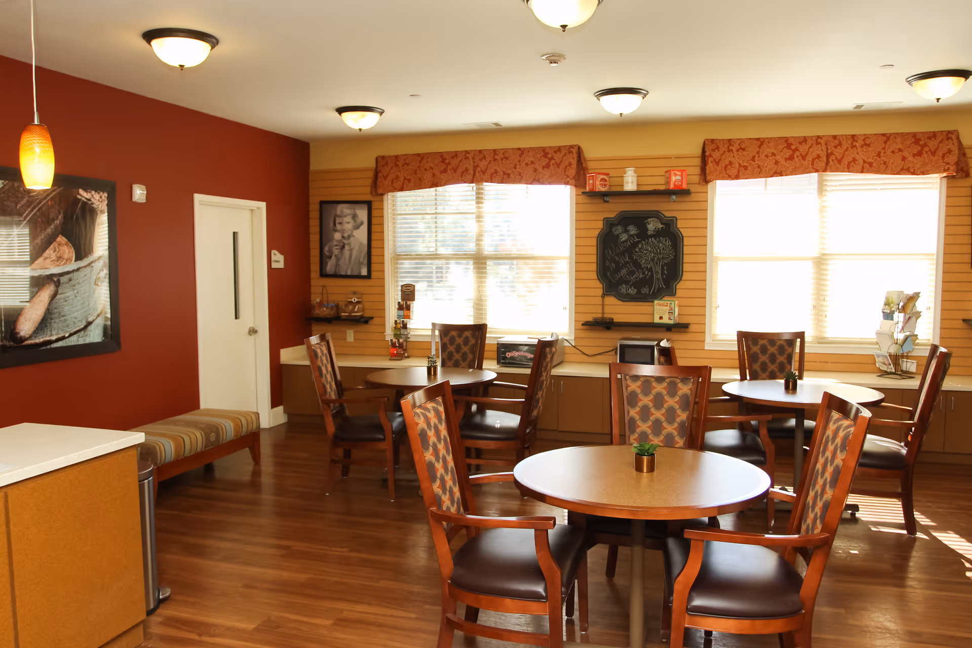 A well-lit dining area with round wooden tables and cushioned chairs featuring patterned upholstery. The room has warm wood flooring, red and beige walls, large windows with valances, and ceiling lights. There is a bench against one wall, framed artwork, and a small chalkboard on the wall near the windows.