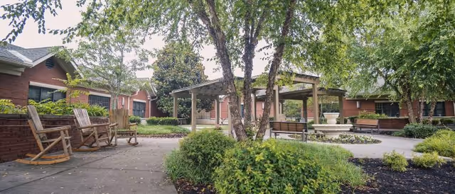 Outdoor courtyard area at Ben Atchley Tennessee State Veterans' Homes featuring a paved walkway, wooden rocking chairs, benches, a pergola, trees, and landscaped greenery with brick buildings in the background.