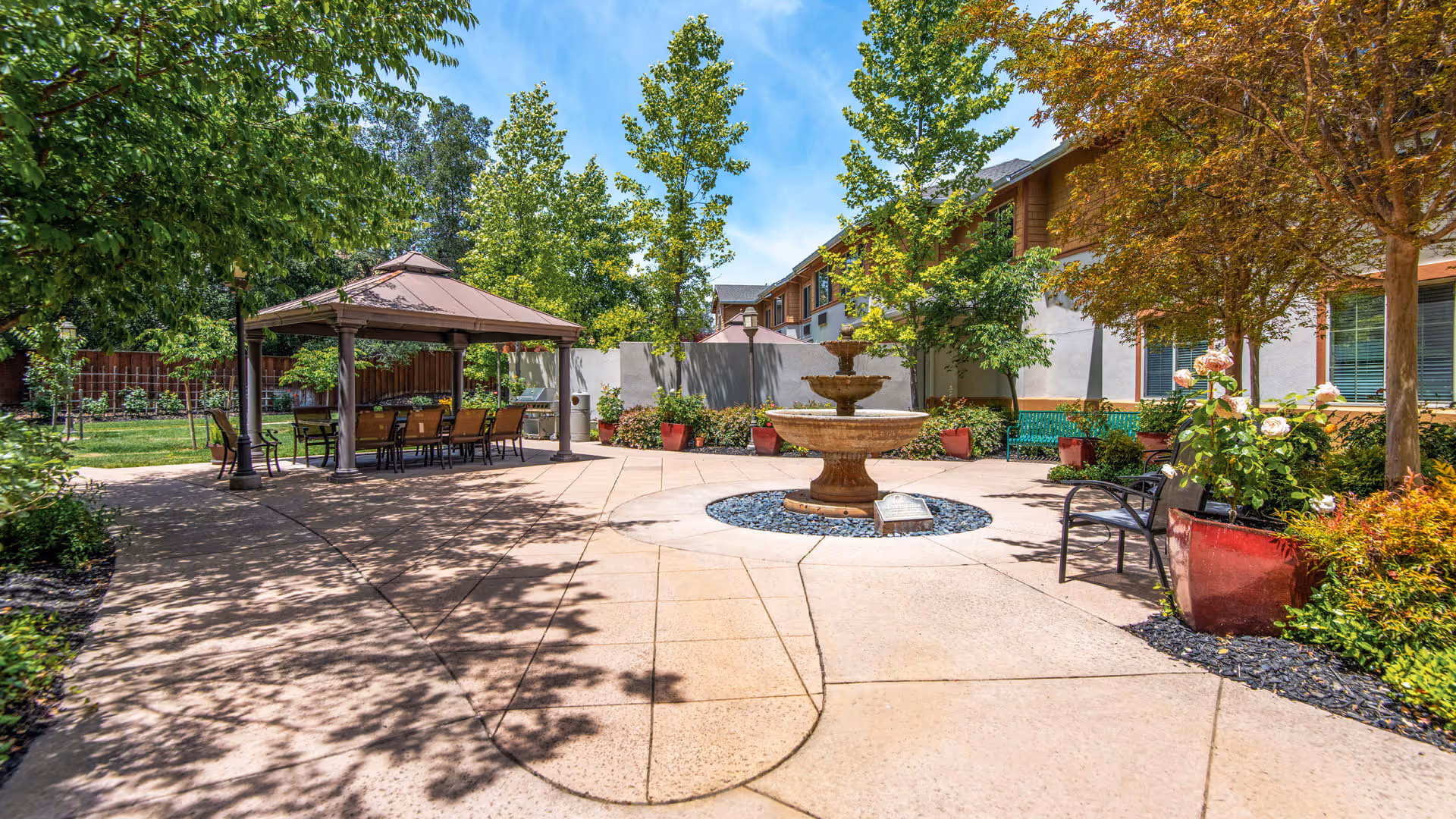 Outdoor courtyard area at Atria Park of Lafayette featuring a central tiered water fountain surrounded by paved walkways, benches, potted plants, and trees. There is a covered seating area with a table and chairs on the left side under a gazebo structure, with a building visible in the background under a clear blue sky.