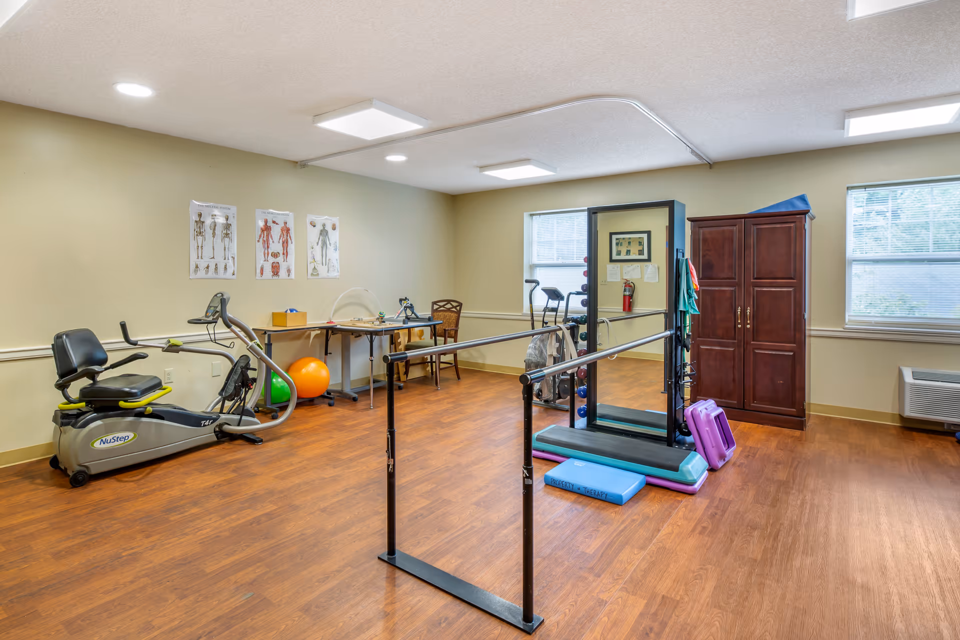 An interior therapy/exercise room with parallel bars, a recumbent exercise bike, mirror, therapy mats and equipment, and a wooden cabinet.