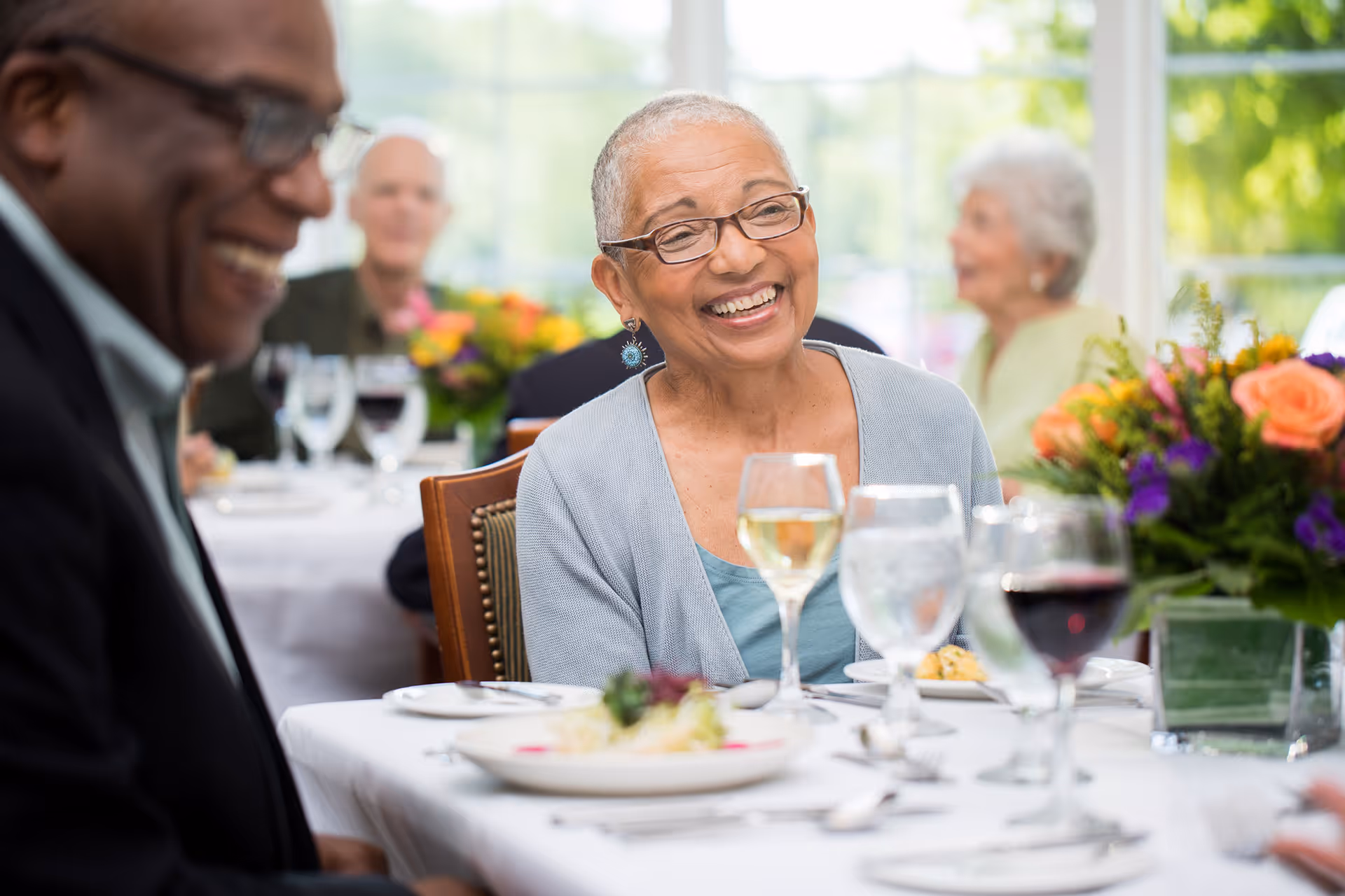 Smiling senior woman wearing glasses seated at a dining table with wine glasses and floral centerpieces, other seniors visible in the background.