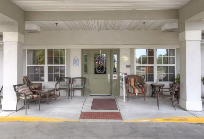 Covered entrance area of a senior living facility with a green double door decorated with a wreath. There are multiple chairs and small tables arranged on both sides of the entrance, some with striped cushions. The area is sheltered by a roof supported by columns, and windows flank the door on either side.