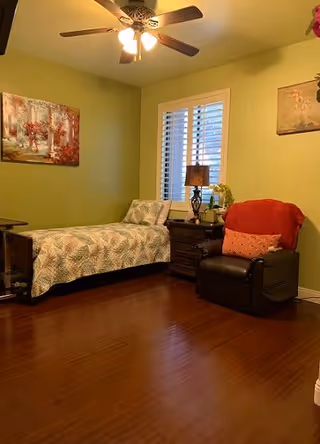 A cozy bedroom with a single bed, nightstand and lamp, recliner chair, ceiling fan, and hardwood floor under green walls.