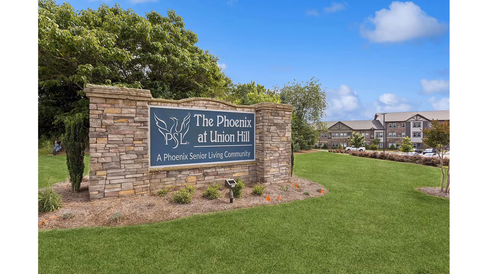 Stone sign for The Phoenix at Union Hill, a Phoenix Senior Living Community, set on a landscaped grassy area with trees and a senior living building in the background under a blue sky.