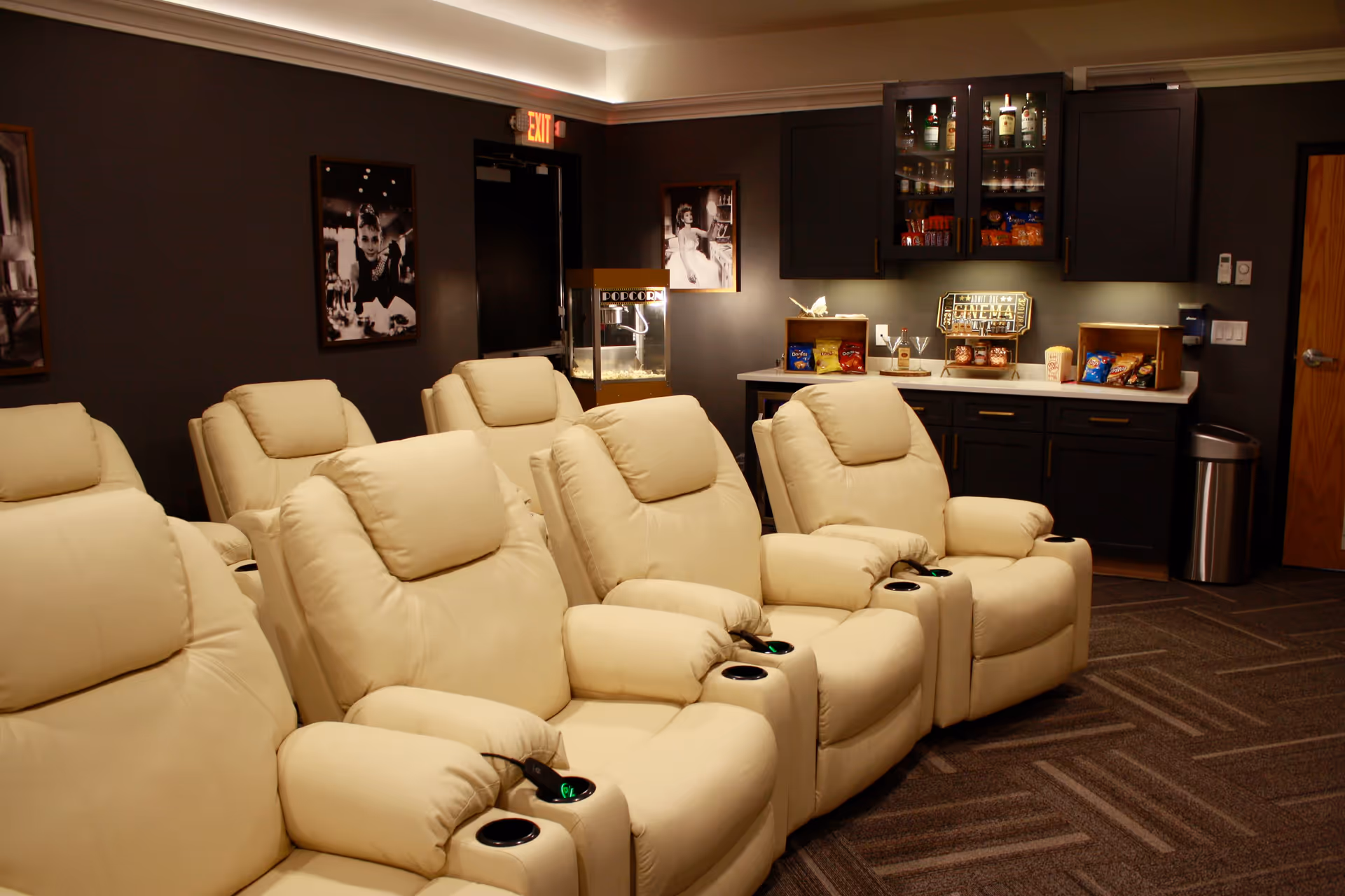 A cozy movie theater room with multiple beige recliner chairs arranged in rows facing a screen. The room has dark walls adorned with black and white classic movie posters. In the back corner, there is a popcorn machine and a snack bar with various snacks and drinks displayed on a countertop and inside cabinets.