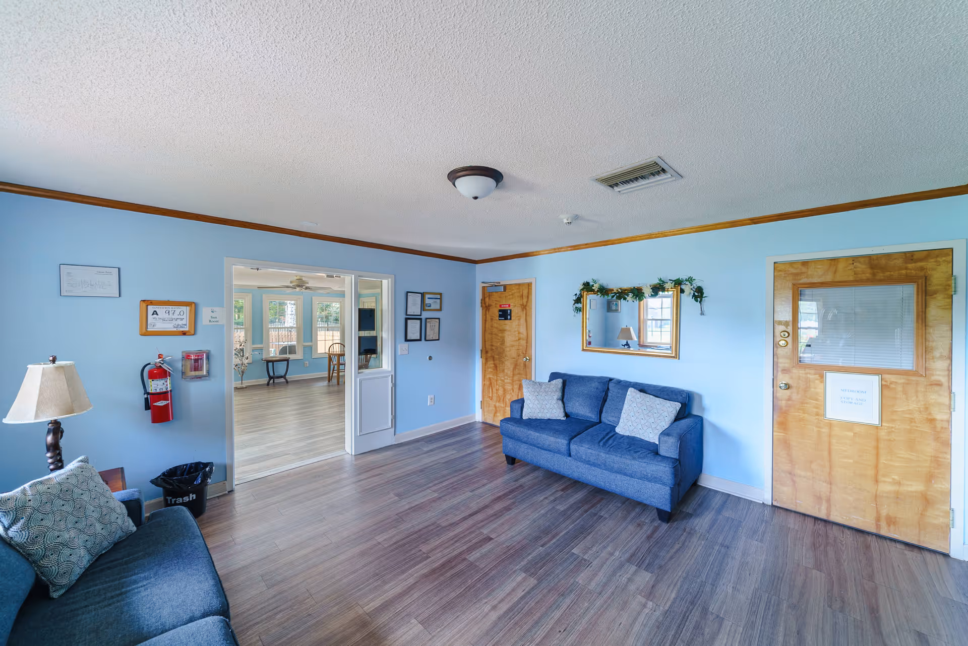 A bright and clean common area with light blue walls and wooden trim. The room features two blue sofas with patterned cushions, a floor lamp, a wall mirror decorated with greenery, and two wooden doors. A fire extinguisher and trash can are visible on the left wall. Through a wide doorway, another room with large windows and wooden flooring is visible.