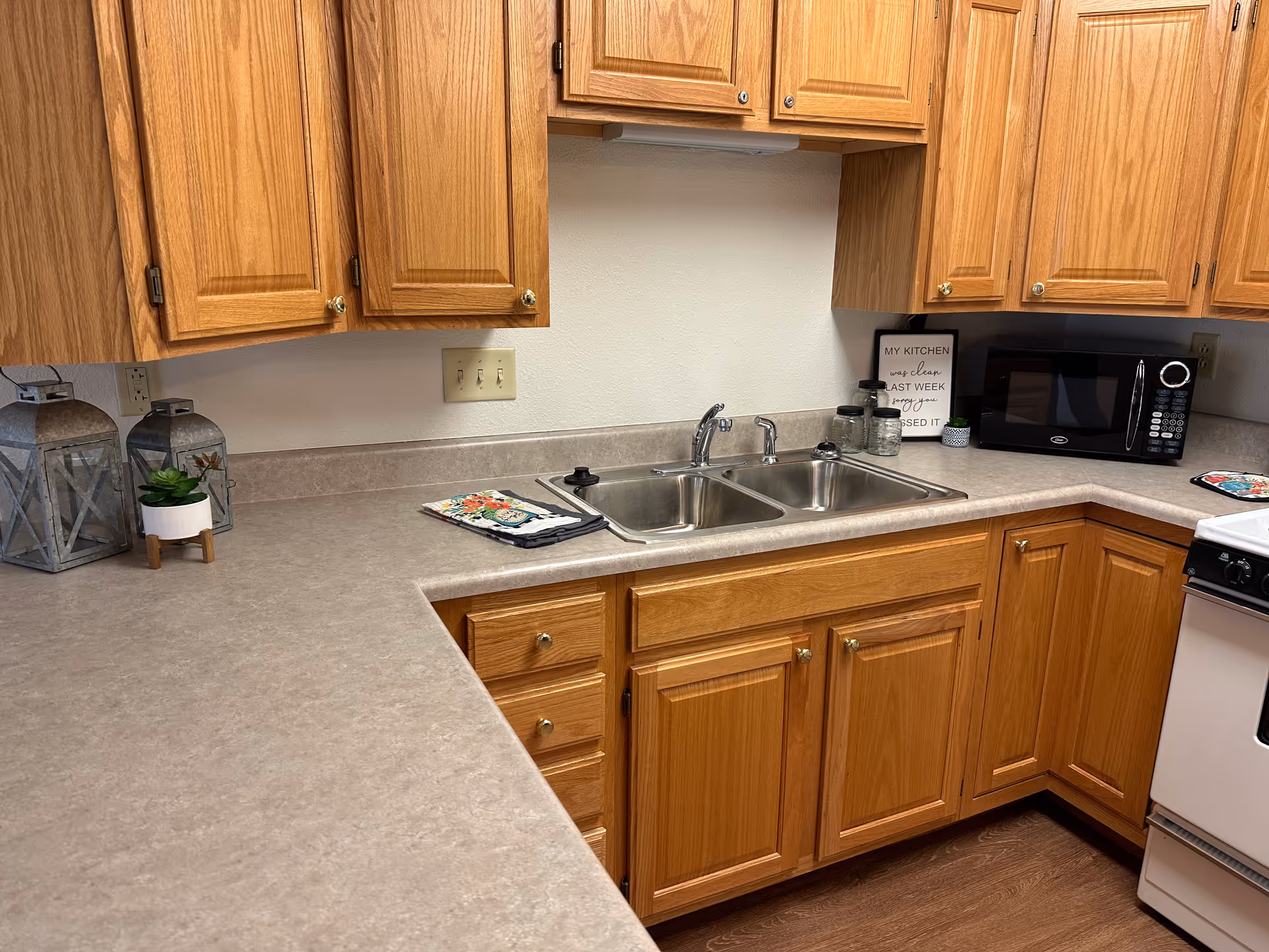 A kitchen with wooden cabinets and a beige countertop. The kitchen features a double stainless steel sink, a black microwave, a white stove, and decorative items including two lanterns and a small potted plant. There is also a sign on the counter that reads, 'MY KITCHEN was clean LAST WEEK sorry you MISSED IT.'