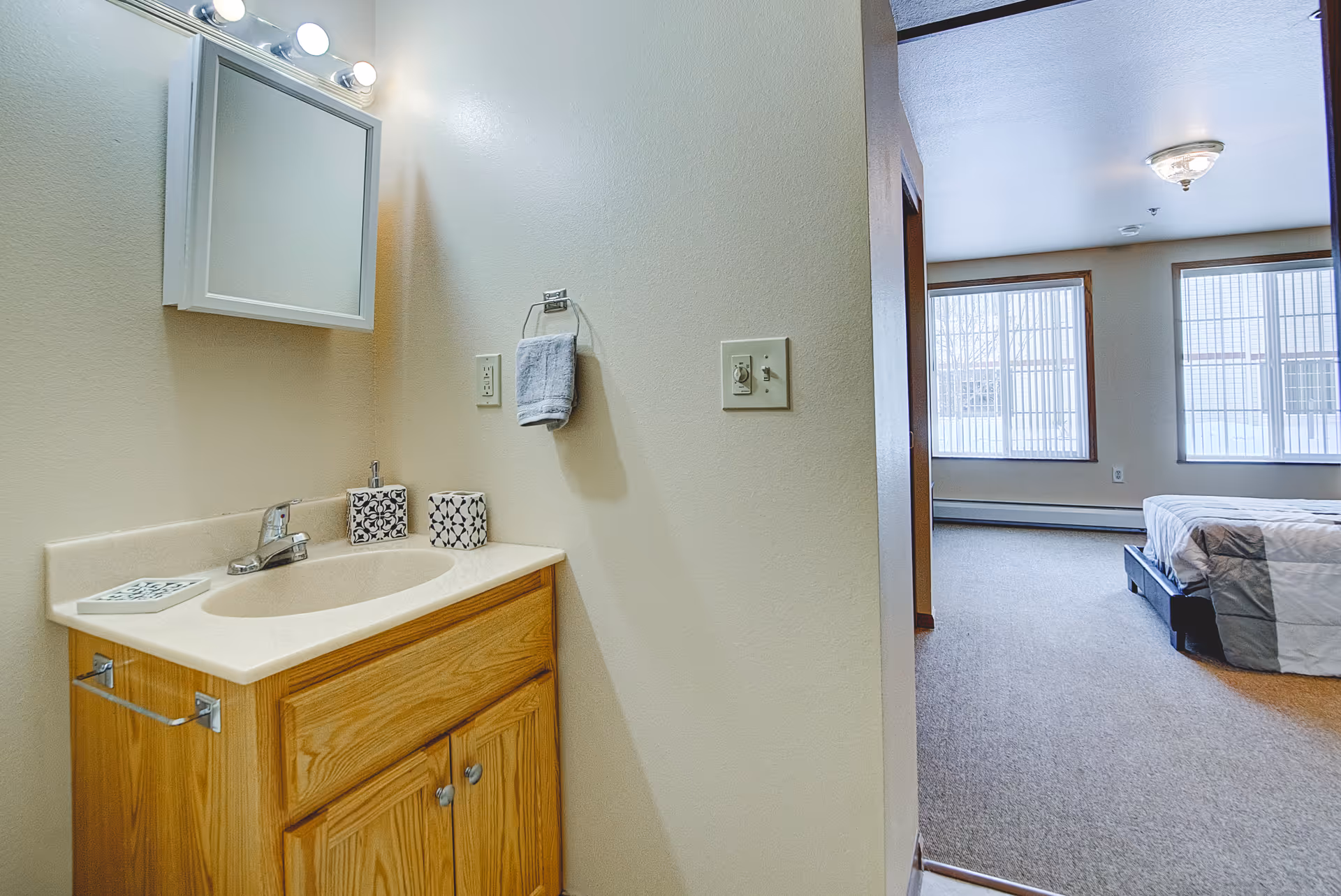 Bathroom vanity and sink beside an open doorway leading to a bedroom with large windows.