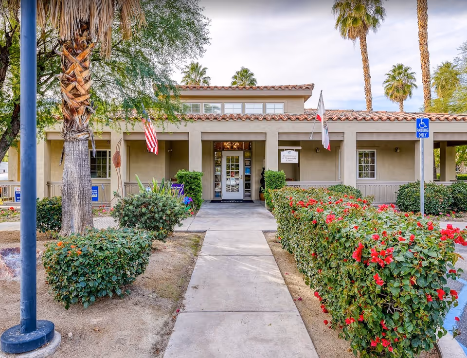 Front exterior view of a single-story building with a tiled roof, surrounded by palm trees and bushes with red flowers. A concrete walkway leads to the entrance, which has a glass door. There are American and California state flags displayed near the entrance, and a sign indicating 'Administration & Community Center'. A blue handicap parking sign is visible on the right side.