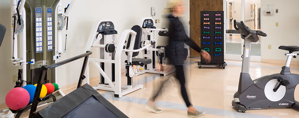 Interior view of a fitness room in a senior living facility with exercise equipment including stationary bikes, weight machines, colorful exercise balls, and a rack of small dumbbells. A person is walking through the room, appearing blurred due to motion.