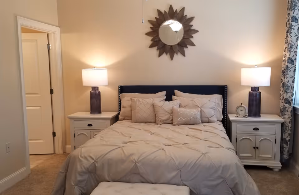 A neatly made bedroom with a bed centered under a decorative sunburst mirror, flanked by matching nightstands and lamps.