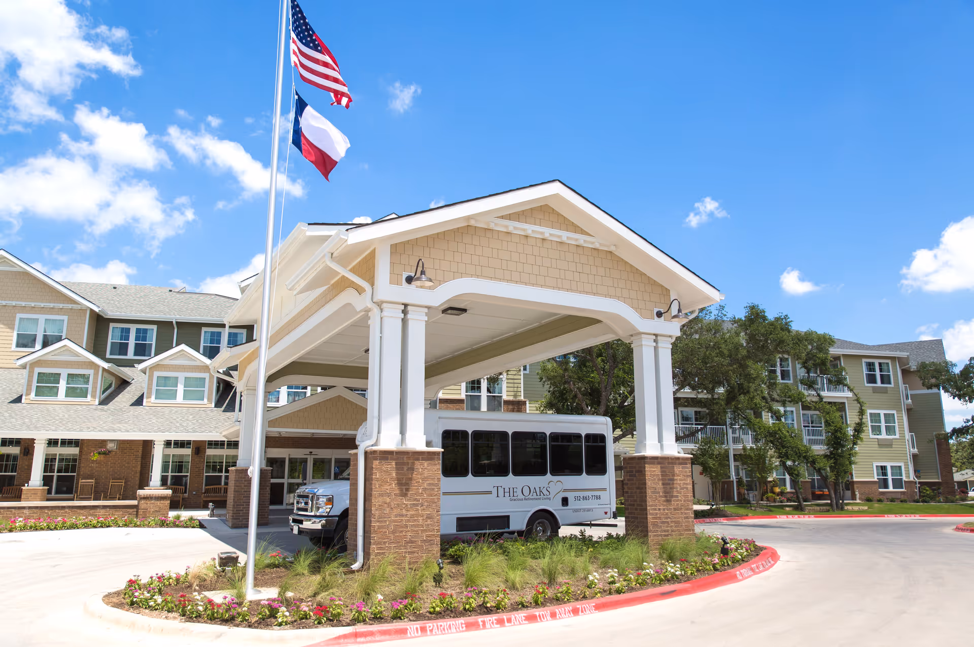 Entrance of The Oaks Gracious Retirement Living facility with a covered drop-off area, a shuttle bus parked under the canopy, two flagpoles with American and Texas flags, and a multi-story residential building in the background under a blue sky with scattered clouds.