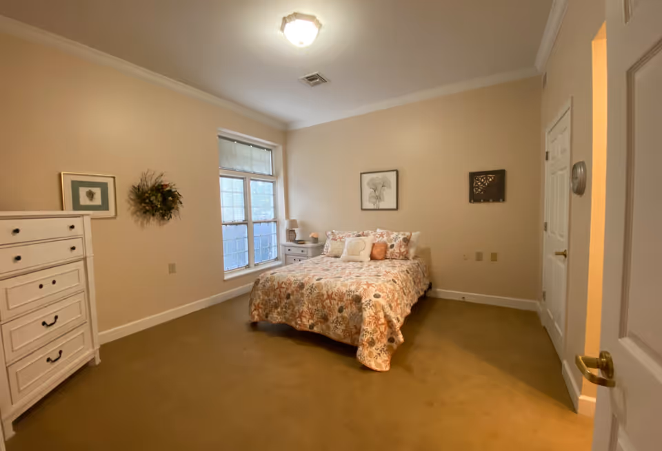 A bedroom with a bed covered in a floral patterned bedspread, two pillows, and a shell-shaped cushion. There is a white chest of drawers on the left, a window with blinds behind the bed, a nightstand with a lamp and decorative items, and framed artwork on the walls. The room has beige walls and carpeted floor.