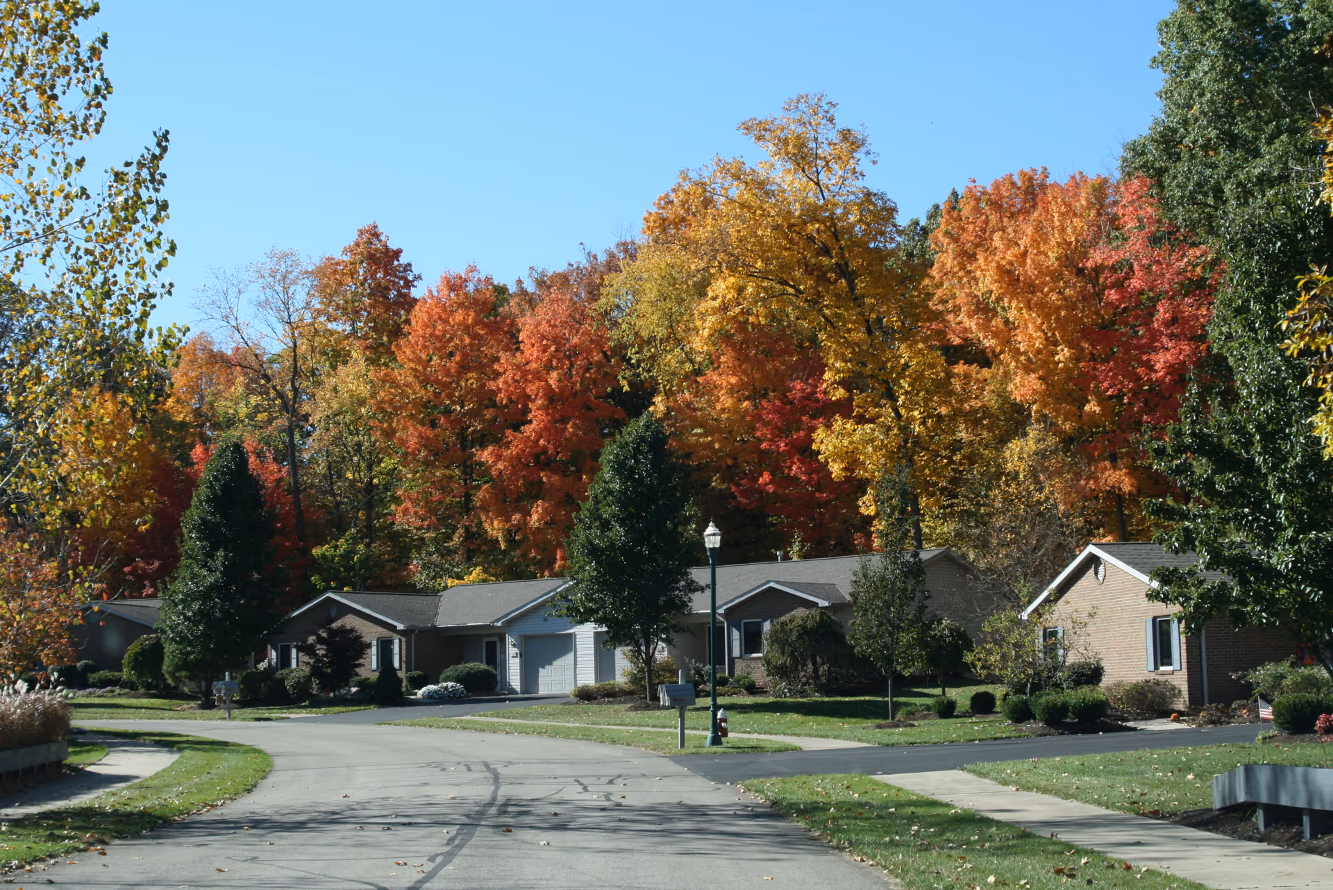A quiet residential street in a senior living community with single-story brick houses surrounded by trees with colorful autumn foliage under a clear blue sky.