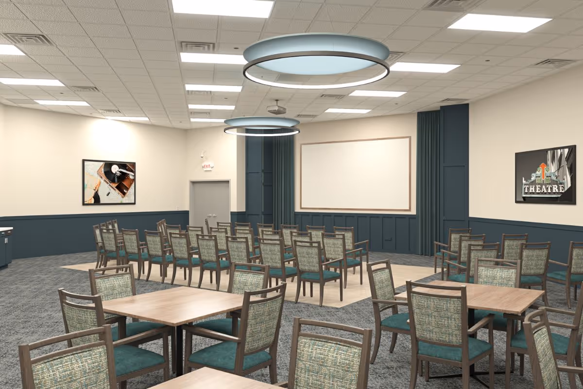 A large meeting or presentation room with rows of chairs facing a blank white screen on the wall. The room has several square tables with chairs around them in the foreground. The walls are painted beige with dark blue wainscoting, and there are two circular ceiling lights. Two framed pictures hang on the walls, one showing a theatre sign and the other a close-up of a turntable.