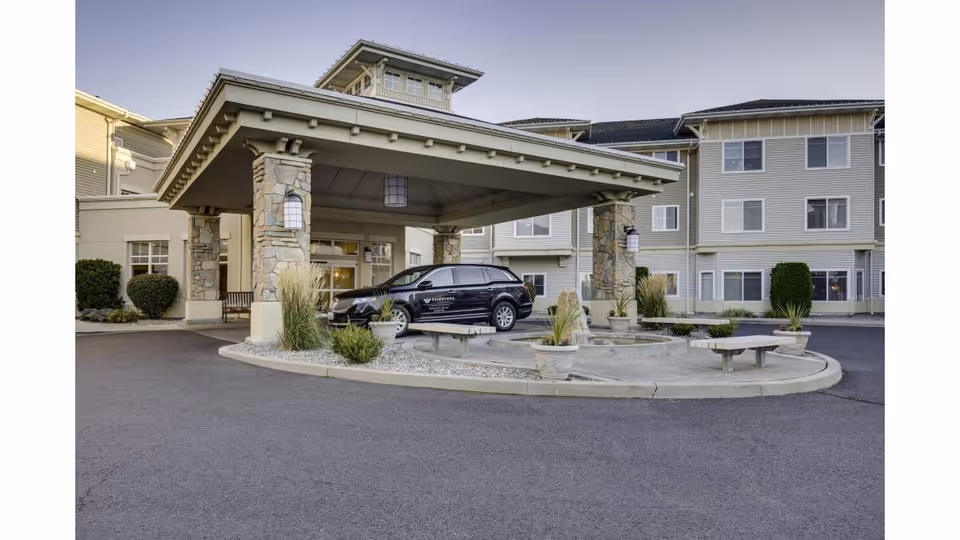 Entrance of a senior living facility with a covered drop-off area supported by stone pillars. A black vehicle is parked under the canopy. The building is a multi-story structure with beige siding and multiple windows. There are landscaped areas with plants and benches near the entrance.