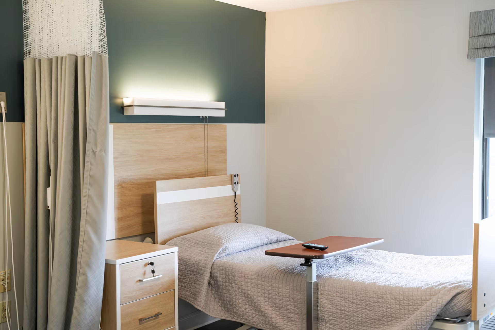 A hospital-style bedroom with a single bed covered in a light gray quilt, a wooden headboard, a bedside table with drawers, a privacy curtain, and a small overbed table with a remote control on it. The wall behind the bed is painted green and white with a modern light fixture above the headboard.