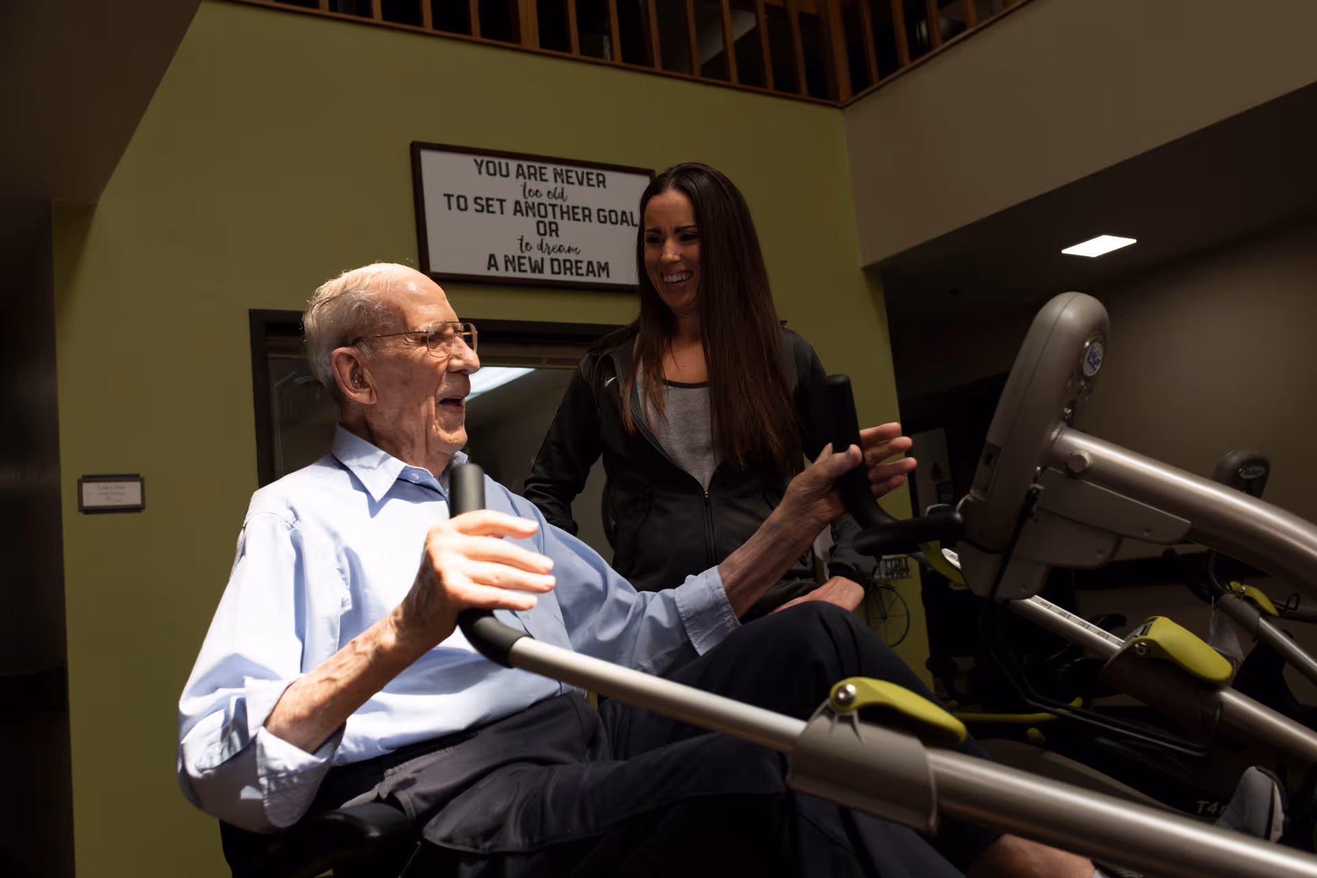 An elderly man is exercising on a seated fitness machine while a younger woman stands beside him, smiling and offering support. They are indoors in a well-lit room with a motivational sign on the wall behind them that reads, 'You are never too old to set another goal or to dream a new dream.'