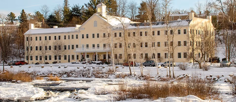 A large, three-story beige building with many windows, set in a snowy landscape with bare trees and a partially frozen body of water in the foreground.