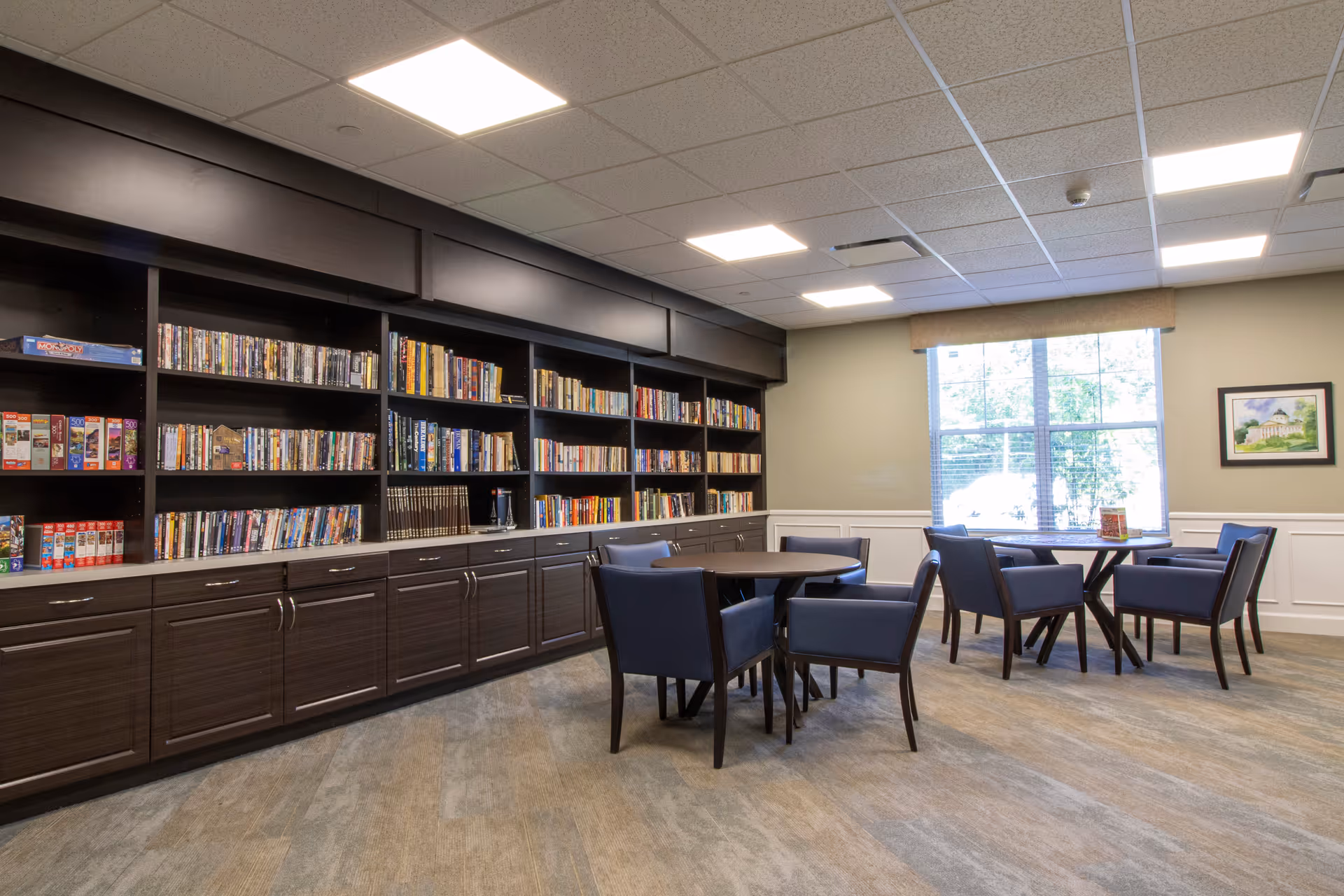 A well-lit room with large dark wood bookshelves filled with books and board games along one wall. In front of the shelves are two round tables, each surrounded by four blue upholstered chairs. A large window with blinds lets in natural light, and a framed picture hangs on the adjacent wall.