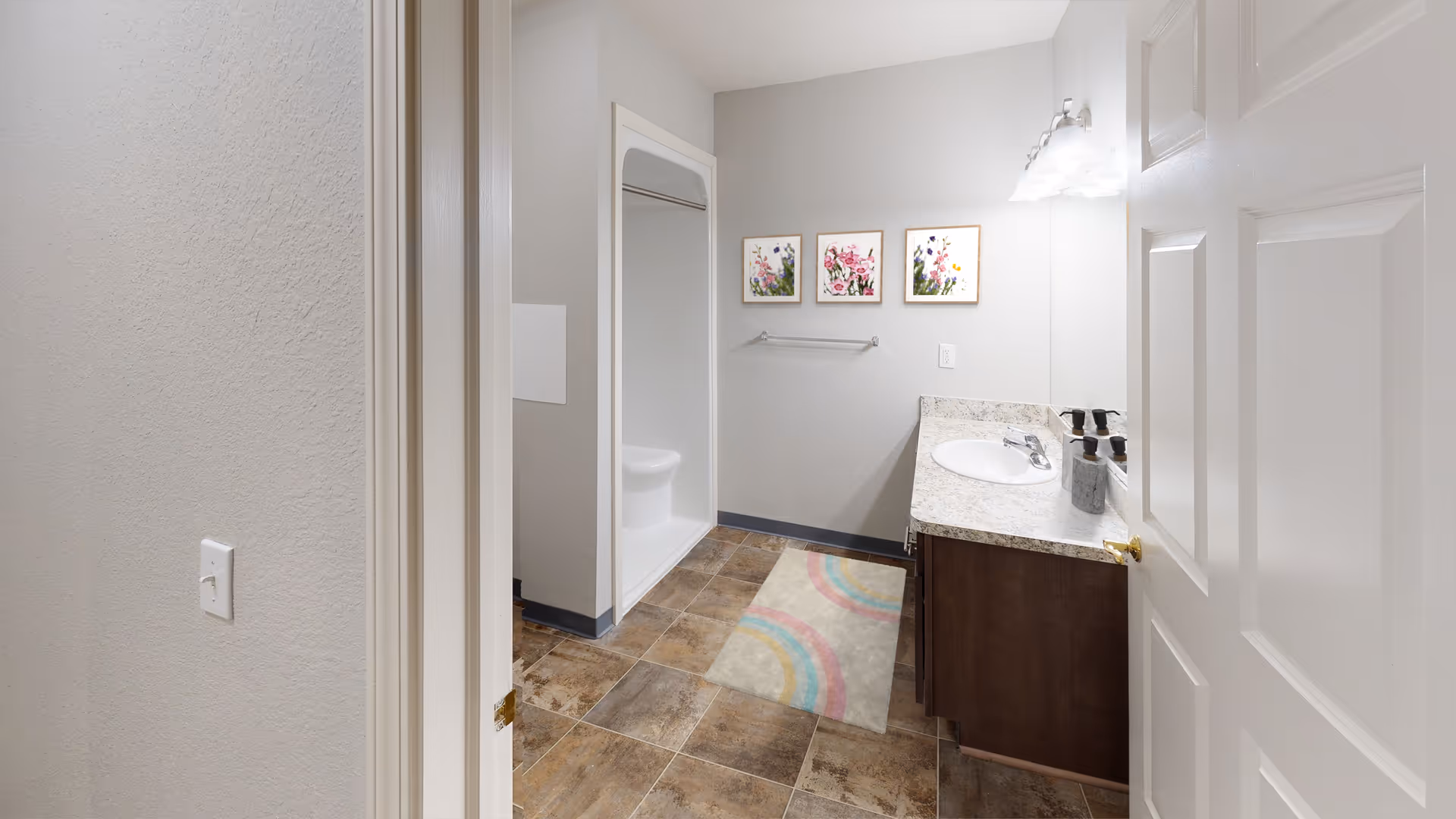 A clean bathroom with a tiled floor, a white shower stall, a countertop with a sink, and three floral paintings on the wall above a towel rack. A colorful rug is placed on the floor in front of the sink.