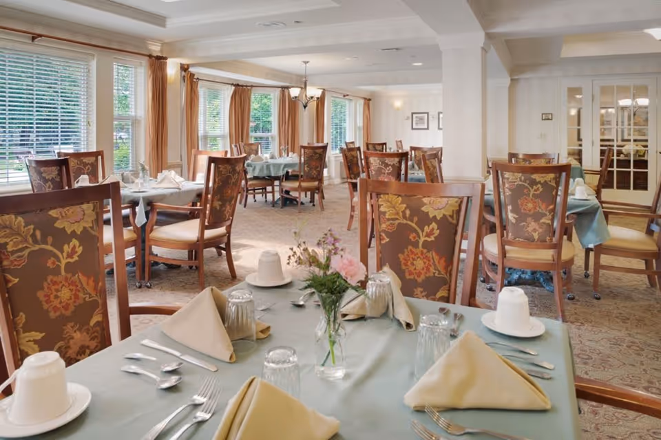 A bright dining room with multiple tables covered in light blue tablecloths, each set with beige napkins, silverware, glasses, and white cups. The chairs have floral patterned upholstery. Large windows with beige curtains allow natural light to fill the room, and a small vase with flowers is placed on the nearest table.