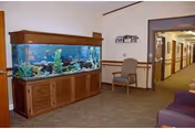 Interior view of a nursing home hallway featuring a large wooden aquarium filled with colorful fish on the left side, a single chair against the wall, and a corridor leading to other rooms. The walls are light-colored with wooden trim, and the carpet is brown.