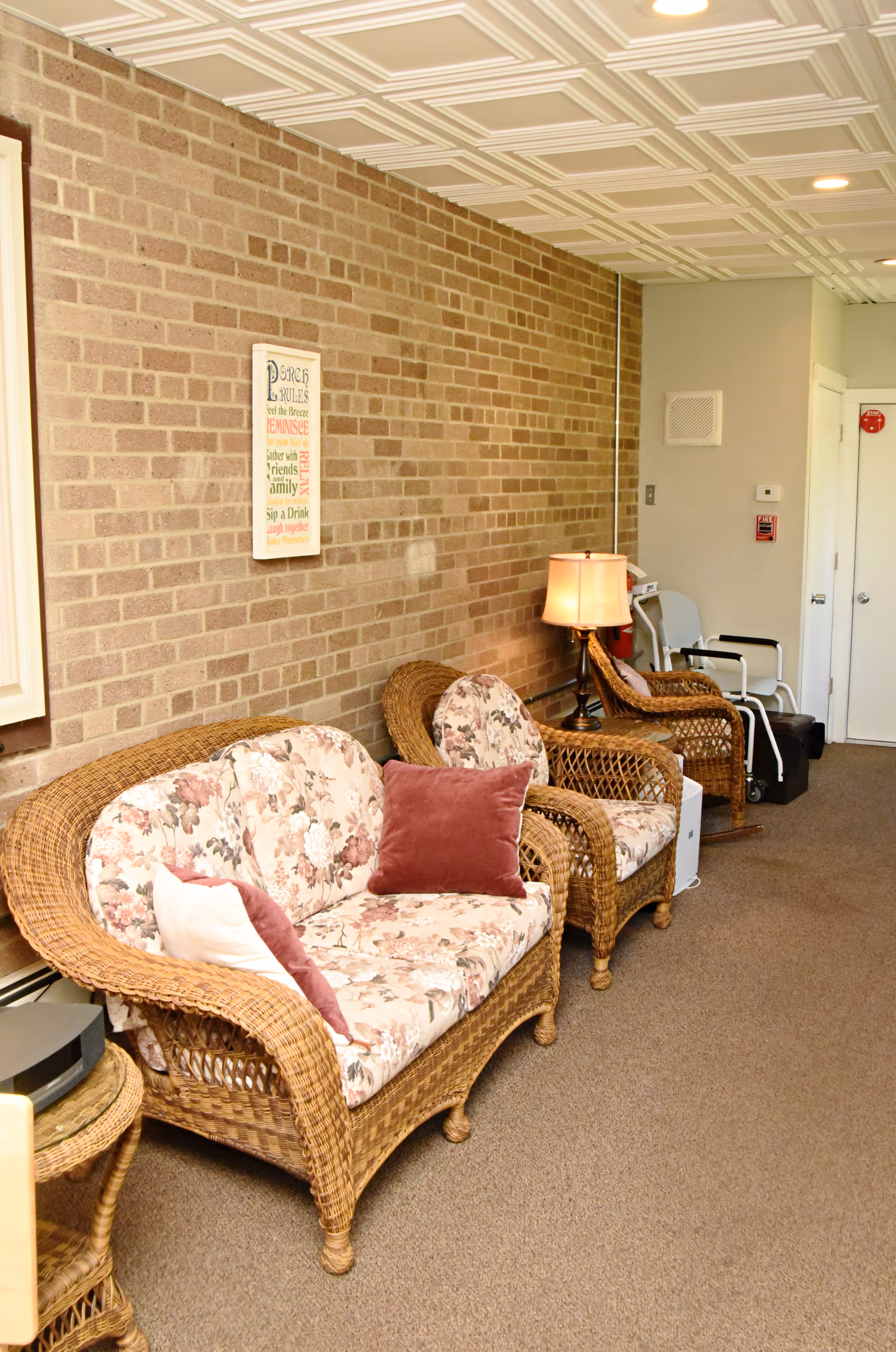 A cozy seating area in a senior living facility with wicker furniture including a loveseat and two armchairs with floral cushions and decorative pillows. A table lamp is placed on a small table between the chairs. The room has a brick wall with a framed inspirational sign, a carpeted floor, and a white paneled ceiling with recessed lighting. A walker and other mobility aids are visible near the back wall.