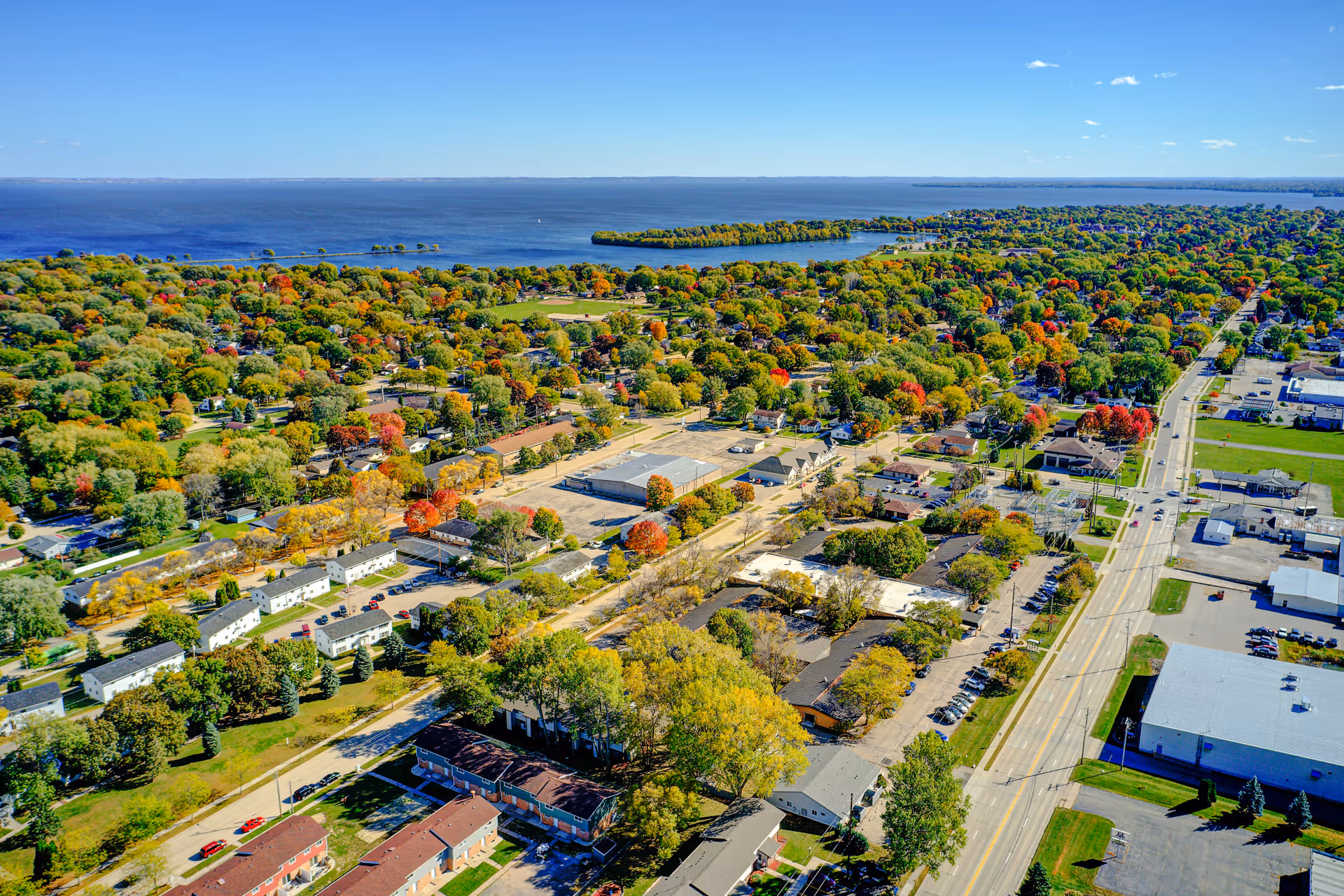 Aerial view of a suburban area with numerous trees showing fall colors, residential buildings, roads, and a large body of water in the background under a clear blue sky.