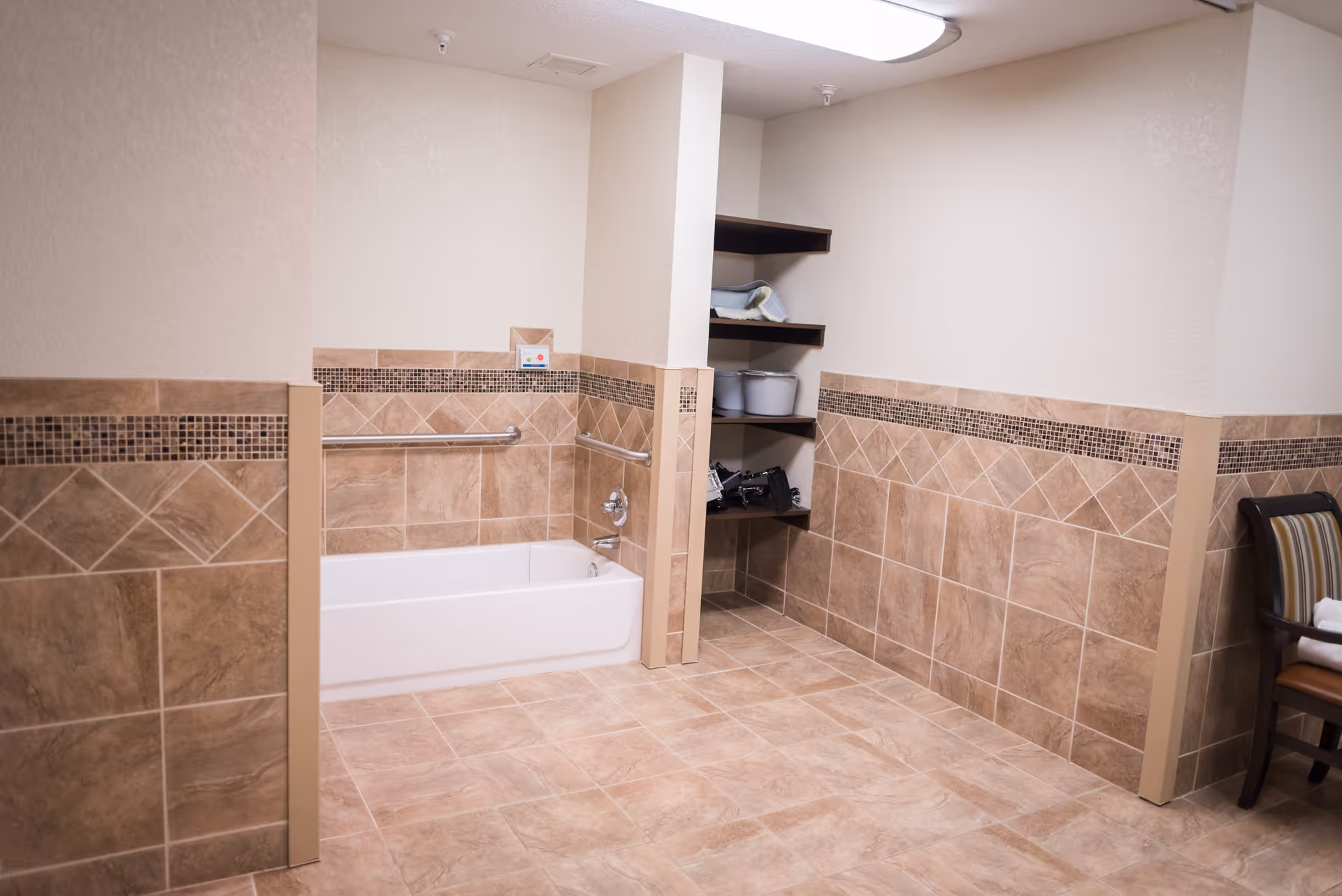 A spacious bathroom with beige tiled walls and floor, featuring a white bathtub with grab bars on the walls for support. There are built-in shelves holding towels and containers, and a chair with a striped cushion is placed to the right.
