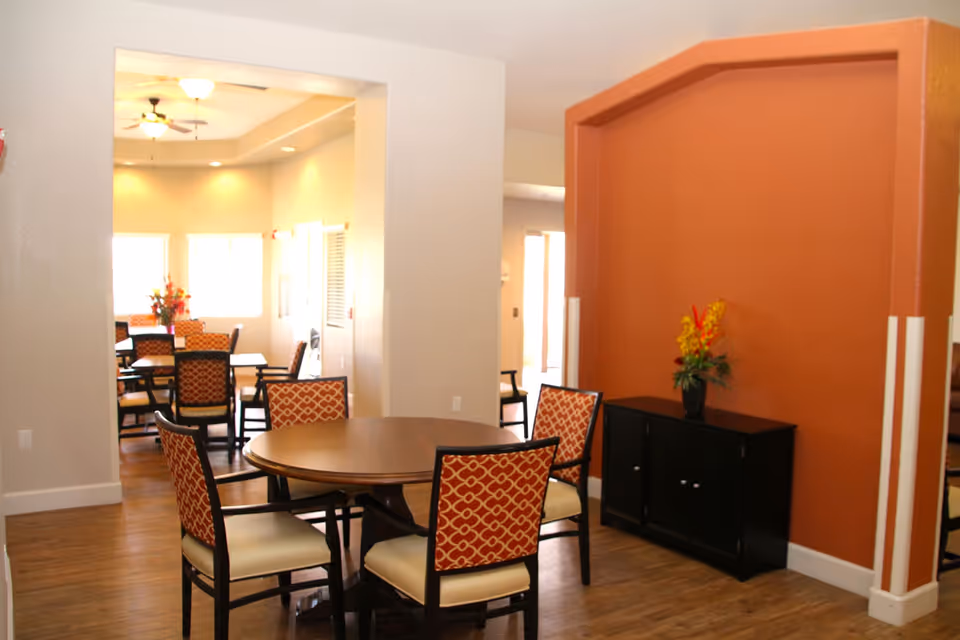 Interior dining room with round wooden tables, patterned chairs, and a side cabinet with flowers against a warm orange accent wall.