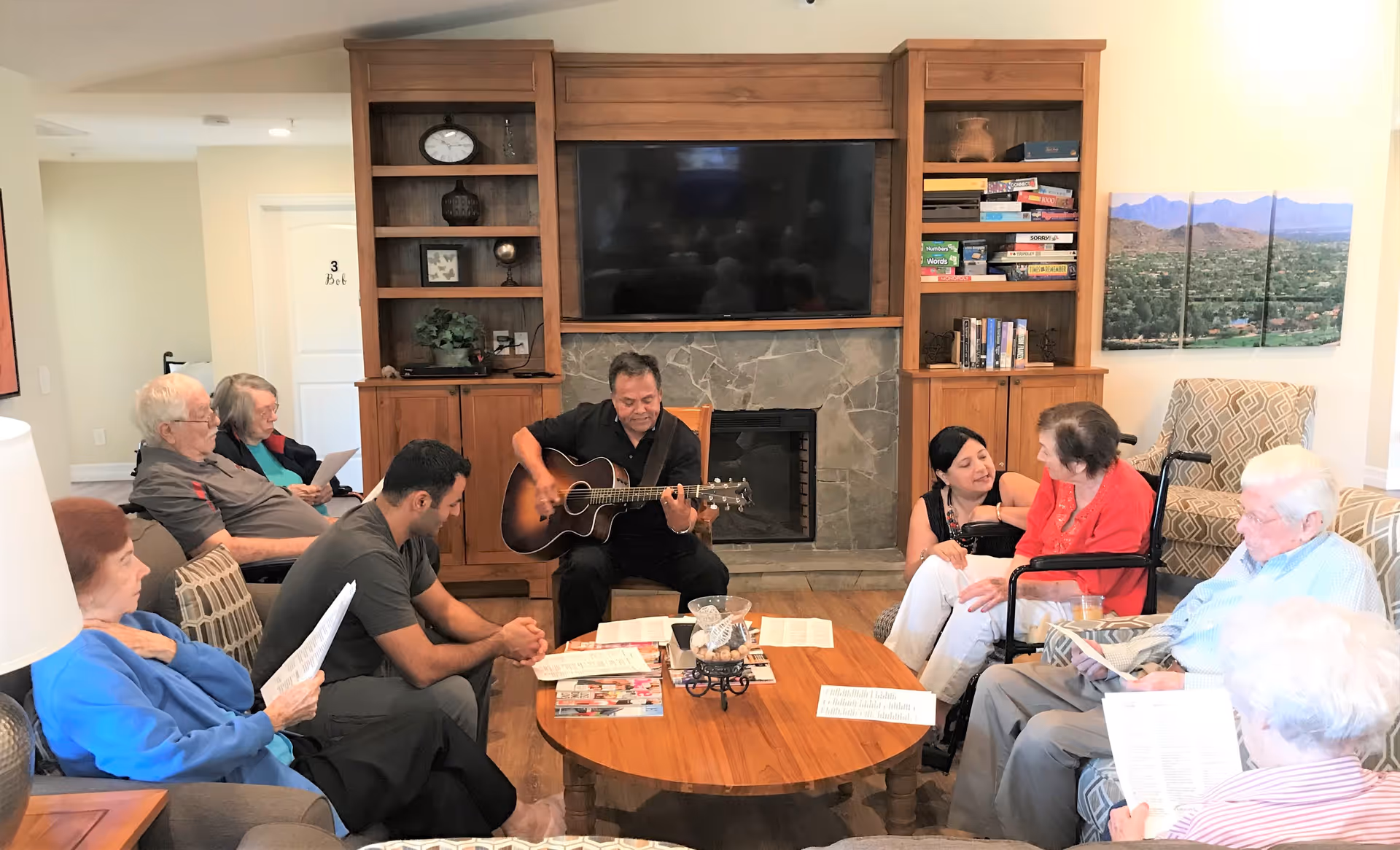 A group of elderly people and two caregivers gathered in a cozy living room. One man is playing an acoustic guitar while others sit around a wooden coffee table, some holding papers, possibly song sheets. The room features wooden built-in shelves with books and board games, a large flat-screen TV above a stone fireplace, and comfortable chairs and sofas.