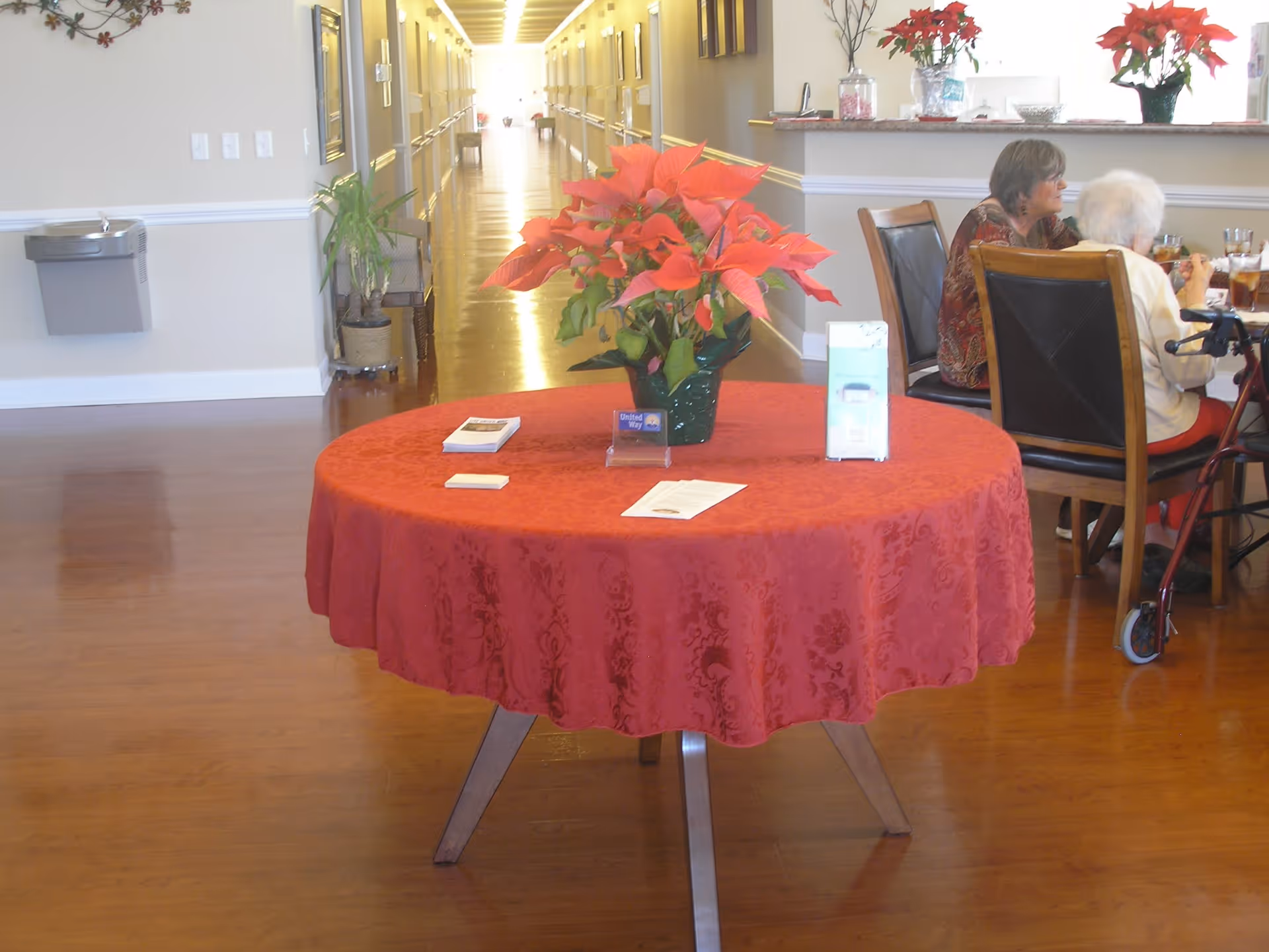 A round table covered with a red tablecloth and a poinsettia plant in a green pot is centered in a senior care facility hallway. On the table are brochures and informational cards. In the background, two elderly women are seated at a dining table, one using a walker. The hallway has wooden floors, light-colored walls, and a water fountain on the left side.