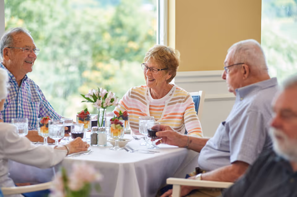 A group of elderly people sitting around a dining table in a well-lit room, enjoying conversation and drinks. The table is set with glasses of water, wine, and fruit parfaits, with a small vase of flowers in the center. Large windows in the background show greenery outside.