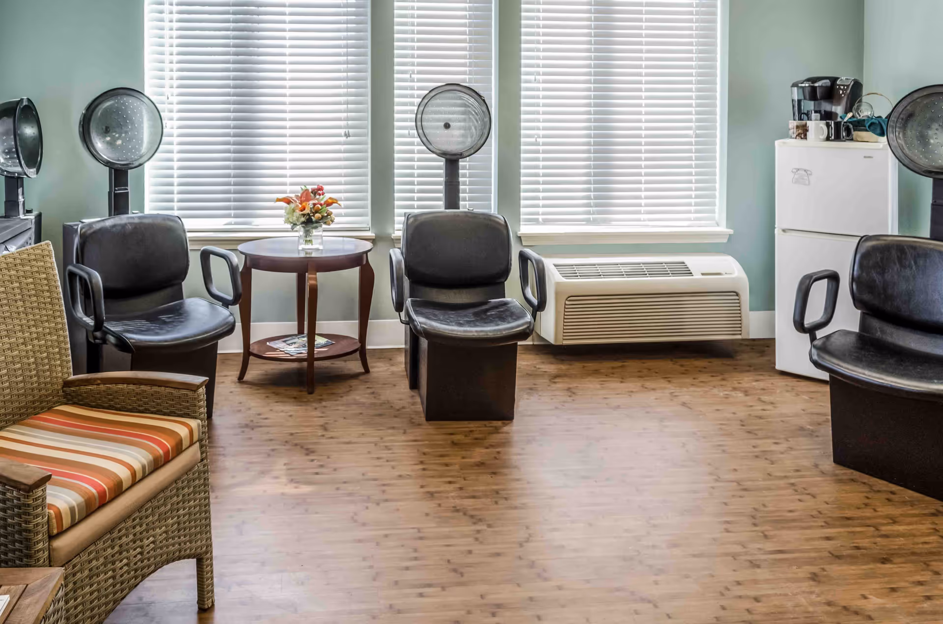 A room with three black salon chairs equipped with hair dryers, a wicker chair with a striped cushion, a small round wooden table with a flower vase, a white mini refrigerator with a coffee maker on top, and large windows with white blinds.