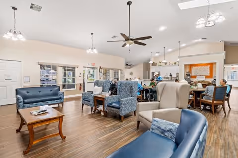 A spacious and well-lit common area in a senior living facility featuring multiple seating options including blue sofas, patterned armchairs, and a beige wingback chair. The room has wooden flooring, ceiling fans, and pendant lights. In the background, several seniors are seated around tables near a kitchen area with a pass-through window.