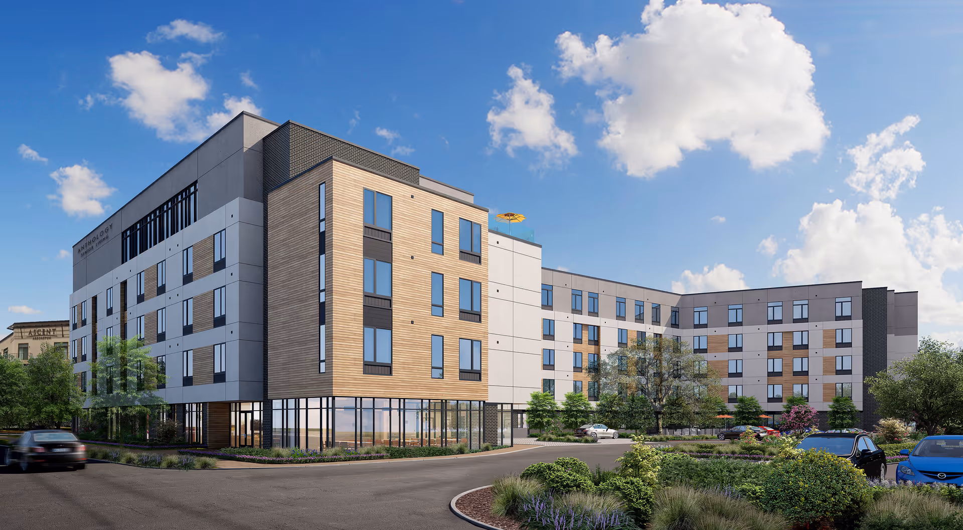 Exterior view of a modern senior living facility building named Anthology Senior Living, featuring multiple floors with large windows, landscaped greenery, parked cars, and a bright blue sky with scattered clouds.