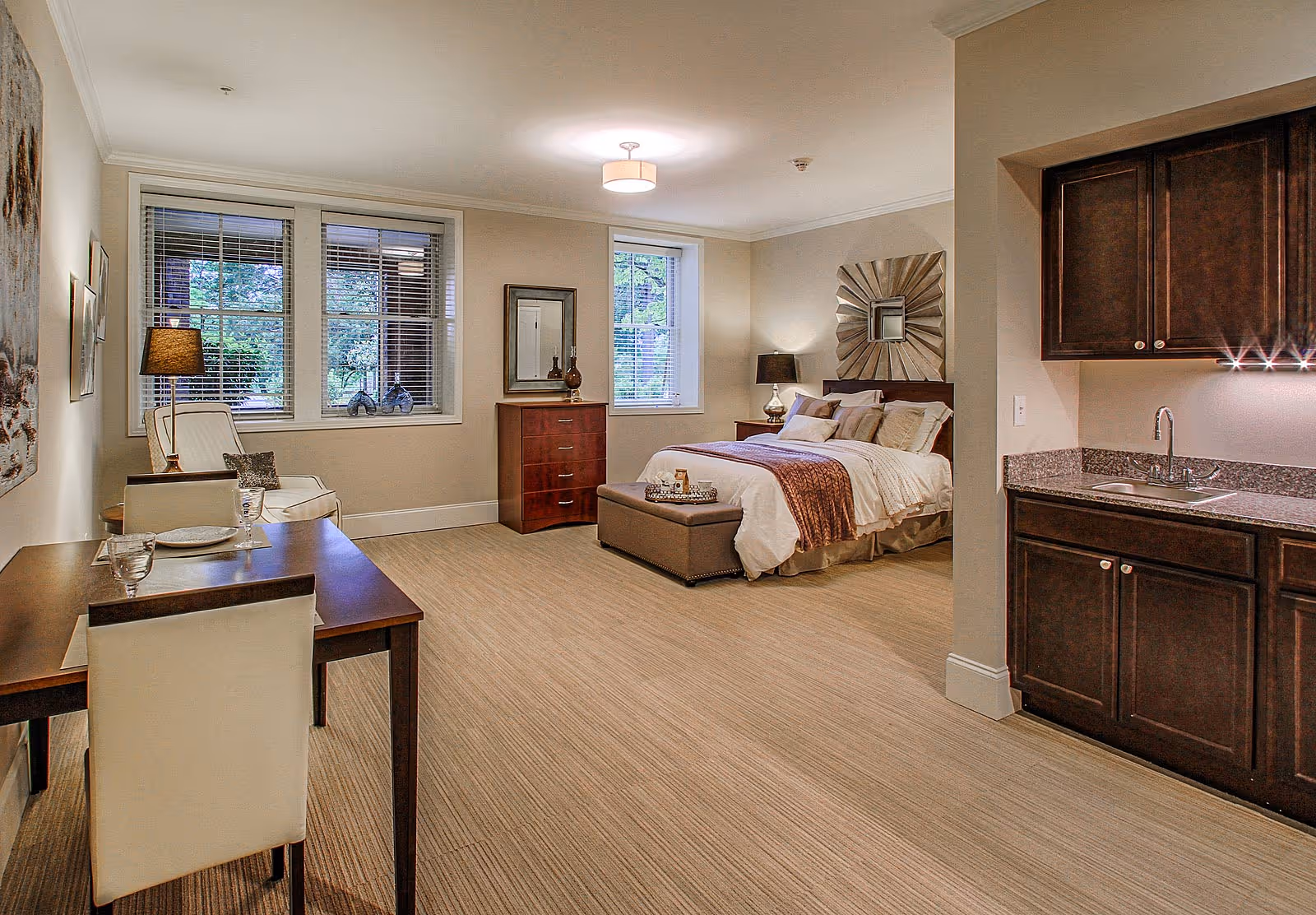 A spacious senior living apartment featuring a bed with pillows and a throw blanket, a cushioned bench at the foot of the bed, a wooden dresser with a mirror, a small dining table with two chairs, a comfortable armchair with a floor lamp beside it, and a kitchenette with dark wood cabinets and a sink. Large windows with blinds allow natural light to enter the room.
