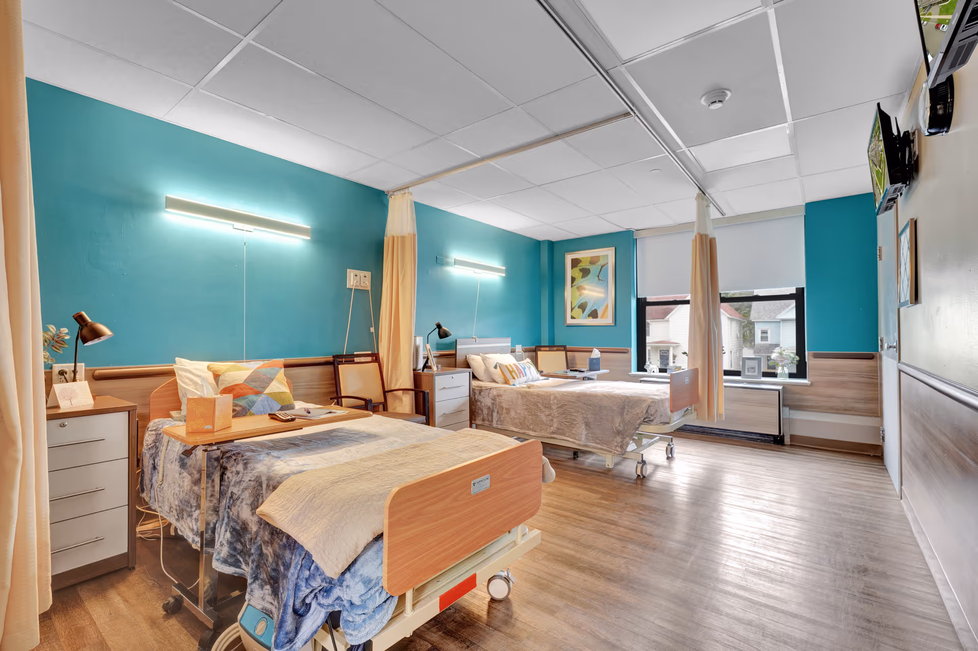 A bright and clean room with two hospital-style beds separated by a curtain. Each bed has colorful pillows and blankets, bedside tables with lamps, and chairs next to them. The walls are painted teal with wood paneling, and there is a window letting in natural light. A television is mounted on the wall opposite the beds.