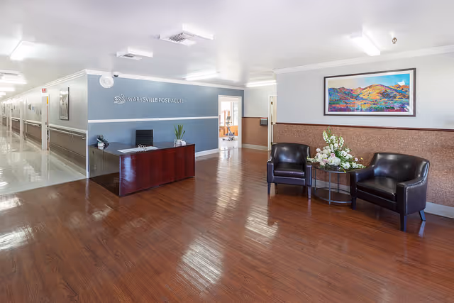 Reception area of Marysville Post-Acute facility with a wooden desk, a chair, and a plant on the desk. To the right, there are two black leather chairs with a small round table between them holding a floral arrangement. A colorful landscape painting hangs on the wall above the chairs. The floor is wooden, and the hallway extends to the left with doors along the corridor.