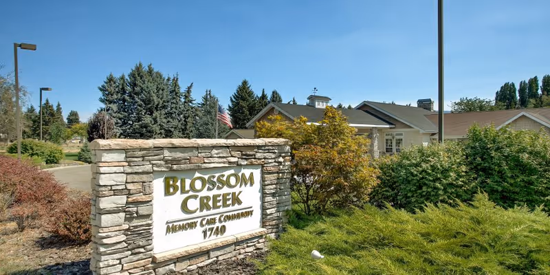 Stone sign for Blossom Creek Memory Care Community with the number 1740, surrounded by bushes and trees, with a building and clear blue sky in the background.