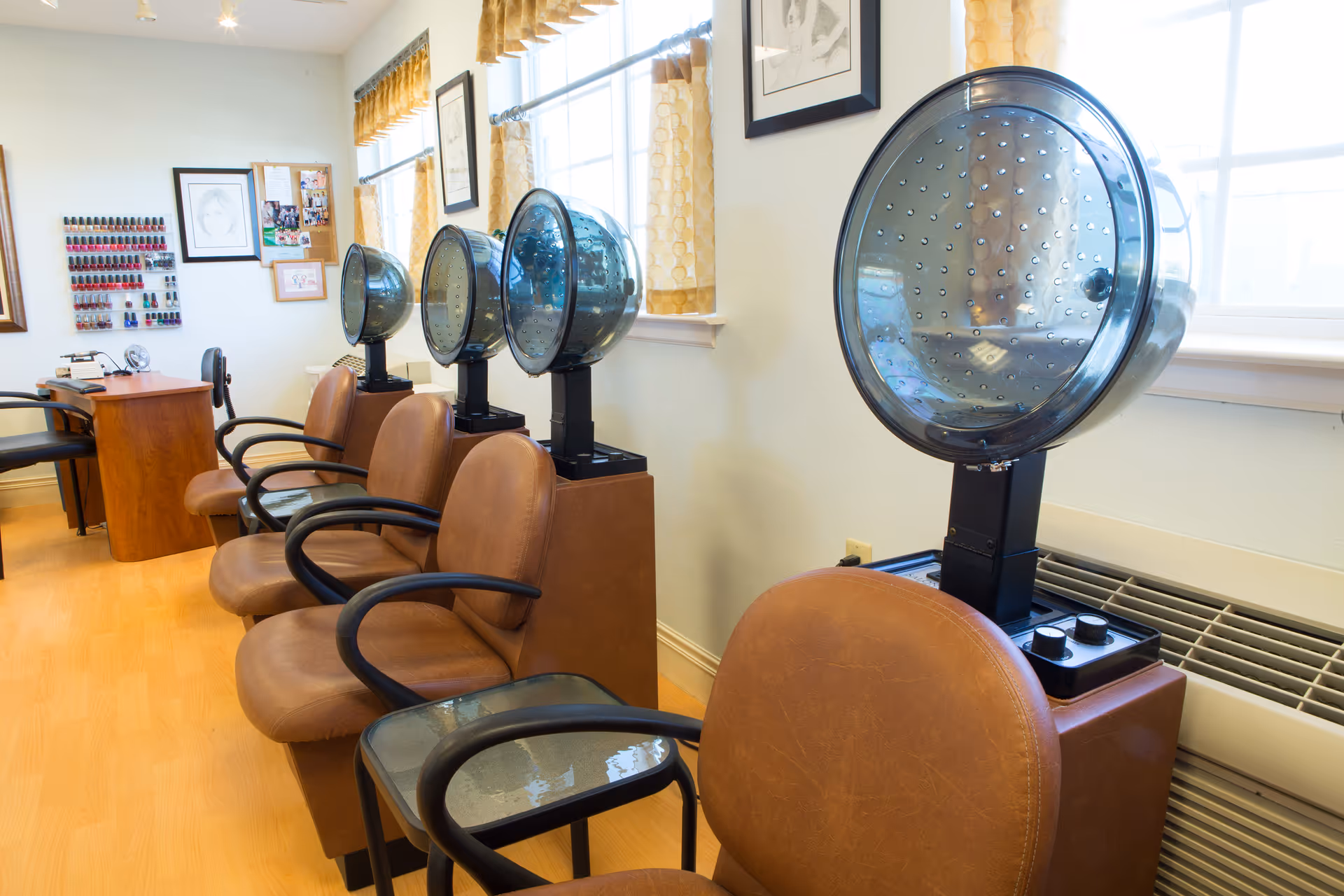 Interior of a salon area with four brown leather chairs each paired with a vintage-style hair dryer hood. The room has light-colored walls, large windows with yellow curtains, a wooden floor, and a desk with a chair in the background. Nail polish racks and framed pictures are visible on the walls.