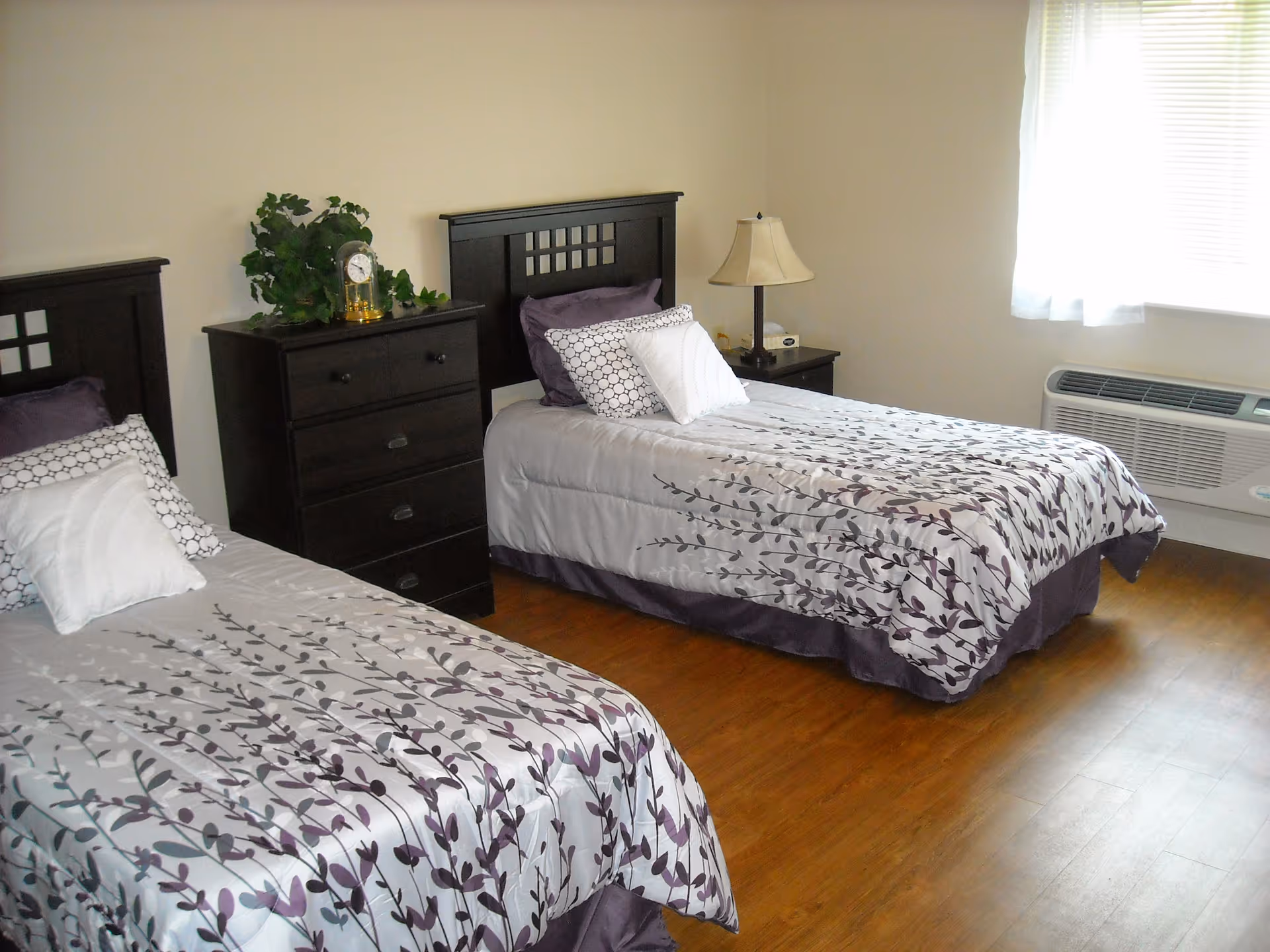 Two-twin-bed bedroom with matching floral bedding, dark wood nightstands and dresser, and a window air conditioner.