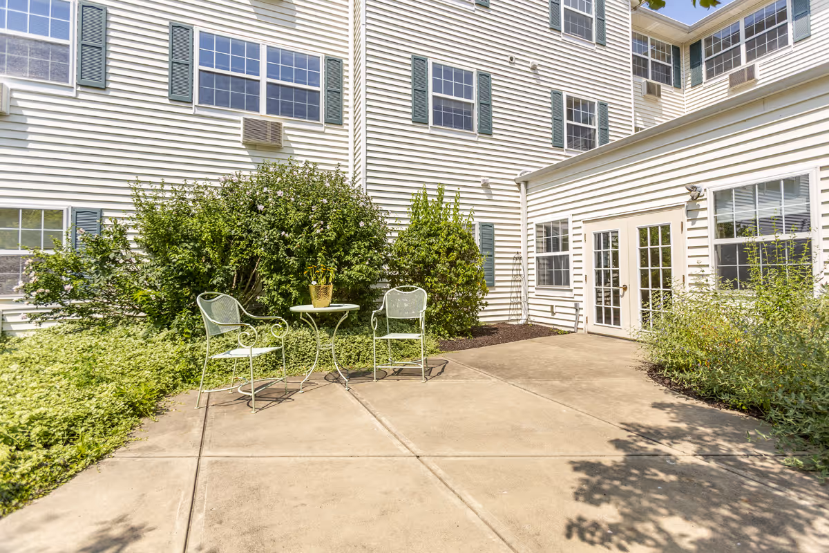 Sunny outdoor courtyard with a small metal table and two chairs in front of a white multi-story building and shrubs.